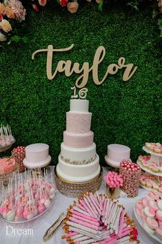 a table topped with lots of cakes and desserts next to a wall covered in greenery