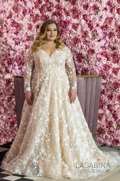 a woman standing in front of a floral wall wearing a long sleeved wedding dress