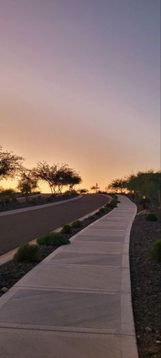 the sun is setting on an empty road with trees and bushes along one side that stretches into the distance