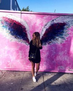 a woman standing in front of a wall with an angel wing painted on it's side