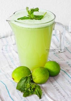 a pitcher filled with green liquid next to three limes and mint leaves on a table