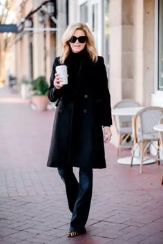 a woman in black coat and sunglasses walking down the street holding a cup of coffee