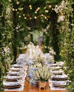 an outdoor dinner table with blue and white dishes, greenery and lights hanging from the ceiling