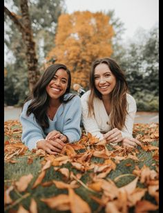 two women laying on the ground with leaves in front of them and smiling at the camera