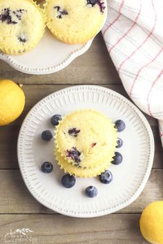 two blueberry muffins on white plates next to lemons and a towel