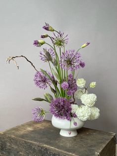 a white vase filled with purple and white flowers on top of a wooden table next to a gray wall