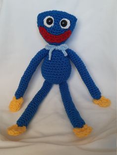 a crocheted blue stuffed animal with yellow feet and legs, sitting on a white sheet