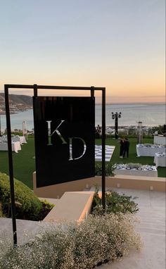 a sign that says k d on it next to some grass and flowers in front of the ocean