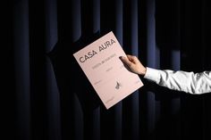 a man holding up a sign that says casa aura on it in front of a black curtain