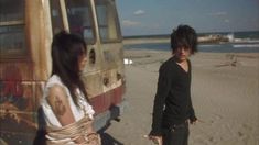 two people standing next to each other on a beach near an old van and the ocean