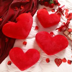 three heart shaped cushions on a bed with red rose petals around them and a gift box in the middle