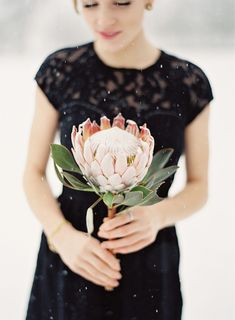 a woman in a black dress holding a pink flower with snow falling on her face