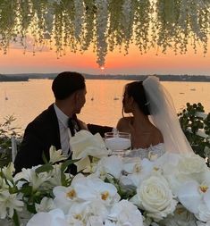 a bride and groom sitting at a table with flowers in front of the sun setting