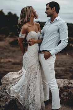 a man and woman standing next to each other on top of a rocky hill with the ocean in the background