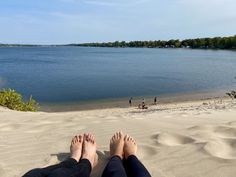 two people are sitting on the sand at the beach looking out over the water and trees