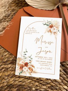 an orange and white floral wedding card on top of a brown envelope next to a fan