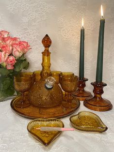 an assortment of candles and dishes on a white table cloth with pink roses in the background