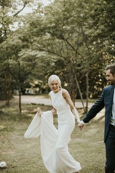 a bride and groom holding hands walking through the grass in front of trees at their wedding