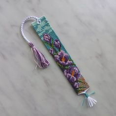 a cross stitch bookmark with tassels and flowers on the front, sitting on a marble surface