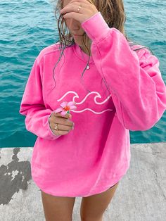 a girl in a pink sweatshirt standing by the water with her hand on her face