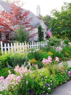 a white picket fence is surrounded by colorful flowers and trees in front of a house