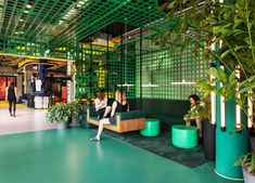 two women sitting on green couches in an indoor space with potted plants and people walking by