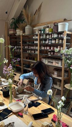 a woman working in a flower shop with flowers on the table and shelves behind her