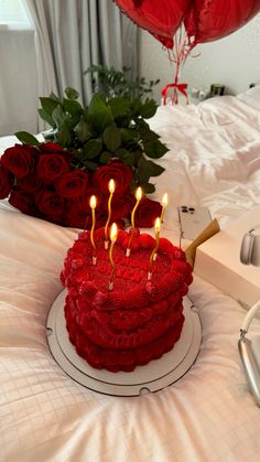 a red cake with candles on it sitting on a bed next to flowers and balloons