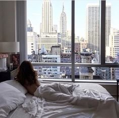 a woman is laying in bed looking out the window at cityscape and skyscrapers