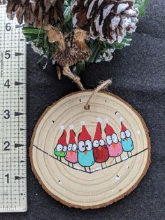 a wood slice with birds painted on it next to pine cones and evergreen needles in front of a ruler