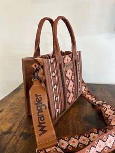 Made of canvas and PU leather trimmed, this tote/crossbody has: Allover Aztec pattern print on canvas Whipstitch details Wrangler logo applique on the front Top zipper closure Inside of the bag includes a zippered pocket and 2 open pockets Double round handle (Drop : 5.75") Detachable/adjustable crossbody strap included 10.5" X 5" X 8.5" (Crossbody strap 46" ) Wrangler Purse, Western Bags, Western Things, Western Bag, Round Handle, Bags Aesthetic, Aztec Pattern, Christmas Wishlist, Crossbody Strap