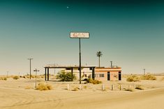 an old gas station in the desert with palm trees and a road sign on top