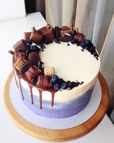 a white cake with chocolate and blueberries on top sitting on a wooden stand next to a window