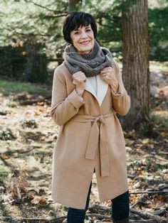 a woman standing in the woods wearing a tan coat and scarf with her hands on her hips