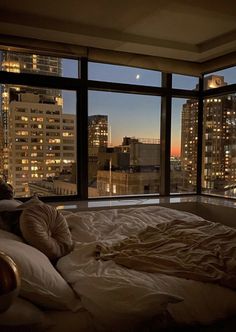 an unmade bed in front of a large window overlooking the city at night time