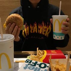 a person sitting at a table with a hamburger and fries in front of him holding a drink