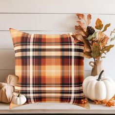 an orange plaid pillow sitting on top of a white shelf next to a vase with leaves and pumpkins