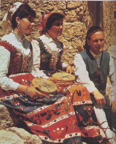 three people dressed in traditional clothing are sitting on some rocks and one woman is holding an instrument