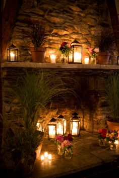 candles are lit in front of a stone fireplace with potted plants and flowers on the mantle