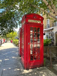 a red telephone booth sitting on the side of a road next to a tree and buildings