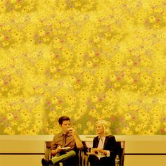 two people are sitting on a bench in front of a wall with yellow and pink flowers