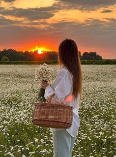 a woman standing in a field holding a basket with flowers on it and the sun setting behind her