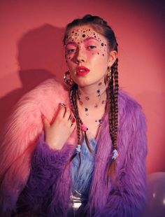 a woman with pink makeup and long braids wearing a purple fur coat, standing in front of a pink wall