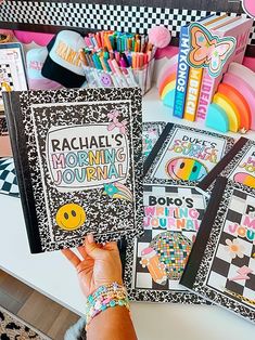 a person holding up a book in front of some other books on a table with pens and pencils