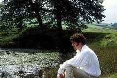 a man sitting on the ground next to a pond with trees in the background and grass around him