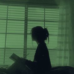 a person sitting on a bed with a book in their hand and the window behind them