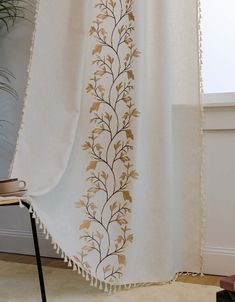 a white and gold embroidered curtain with tassels on the bottom, in front of a window