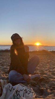 a woman sitting on top of a sandy beach next to the ocean at sun set