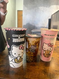 three hello kitty cups sitting on top of a wooden table next to a person holding a cell phone