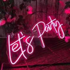 a neon sign that says let's party on the side of a fence with pink flowers